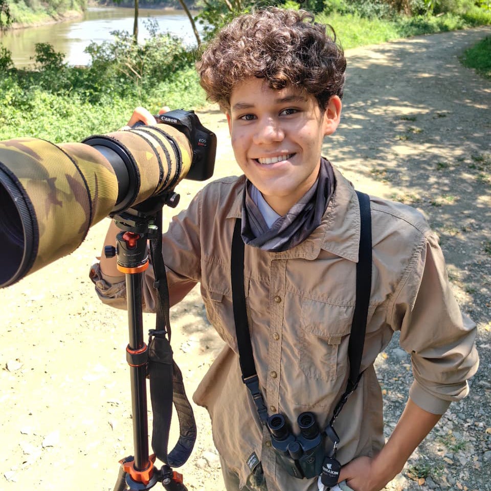 A young person stands in front of a camera with a long lens, binoculars are hanging from his shoulders. 